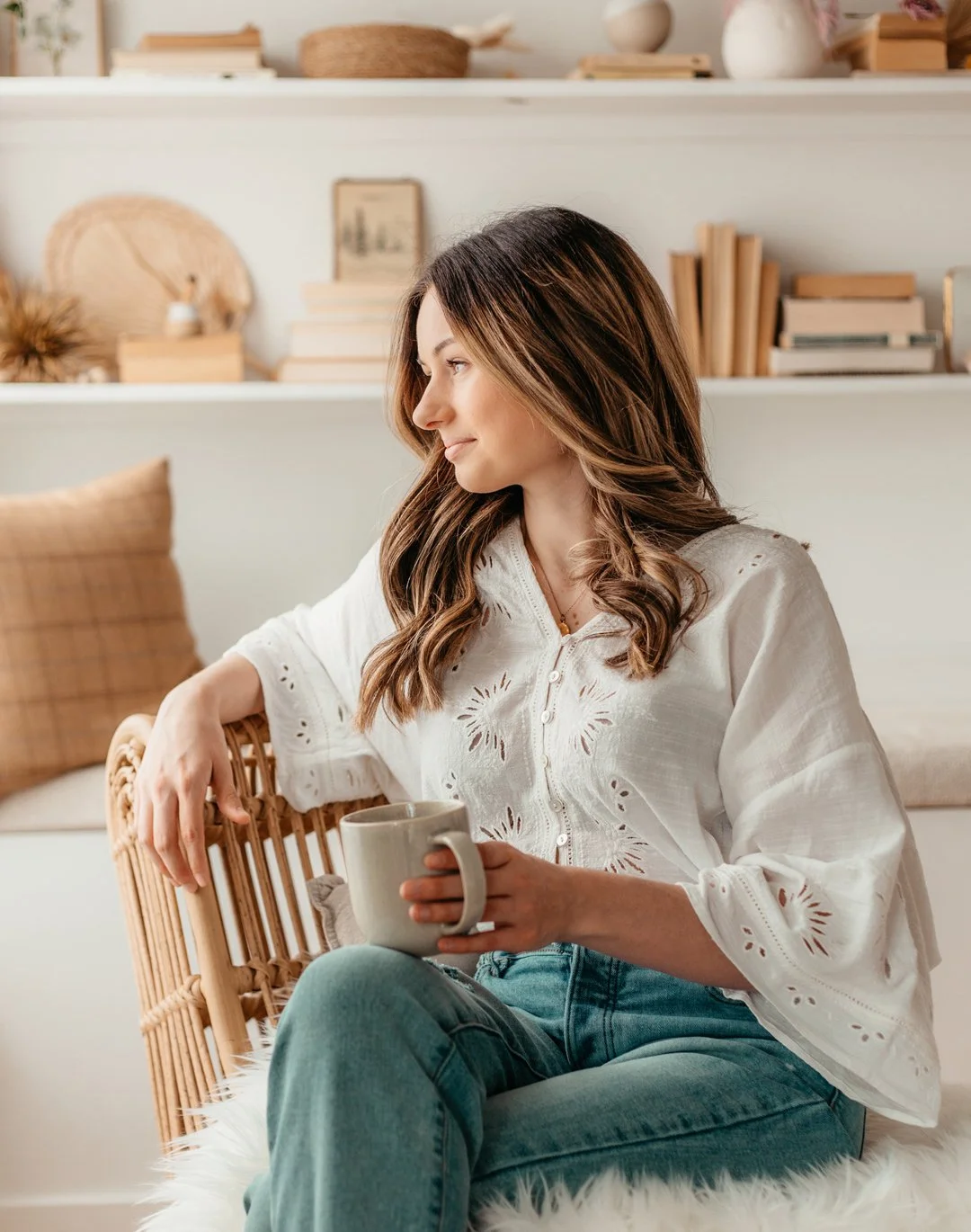 Woman sitting on a rattan chair holding a mug, with a white shirt and blue jeans, in a cozy home setting with shelves of books and decor in the background.