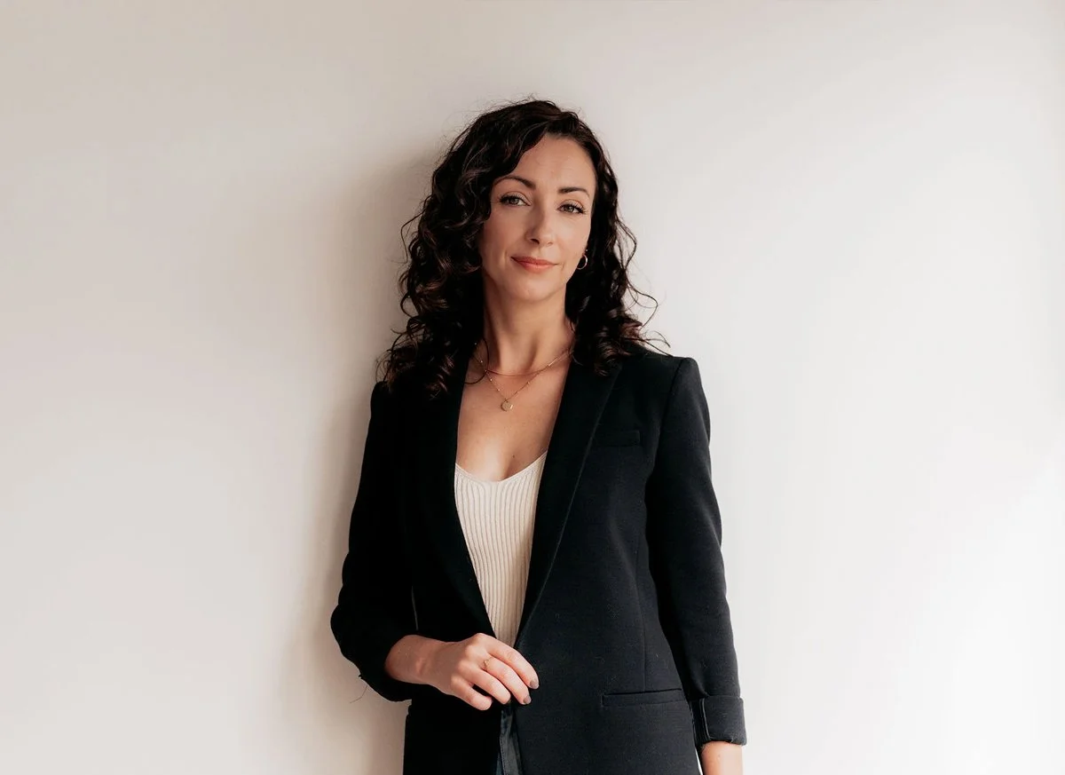 A woman with dark, curly hair wearing a black blazer and a white top, standing against a plain light-colored wall.