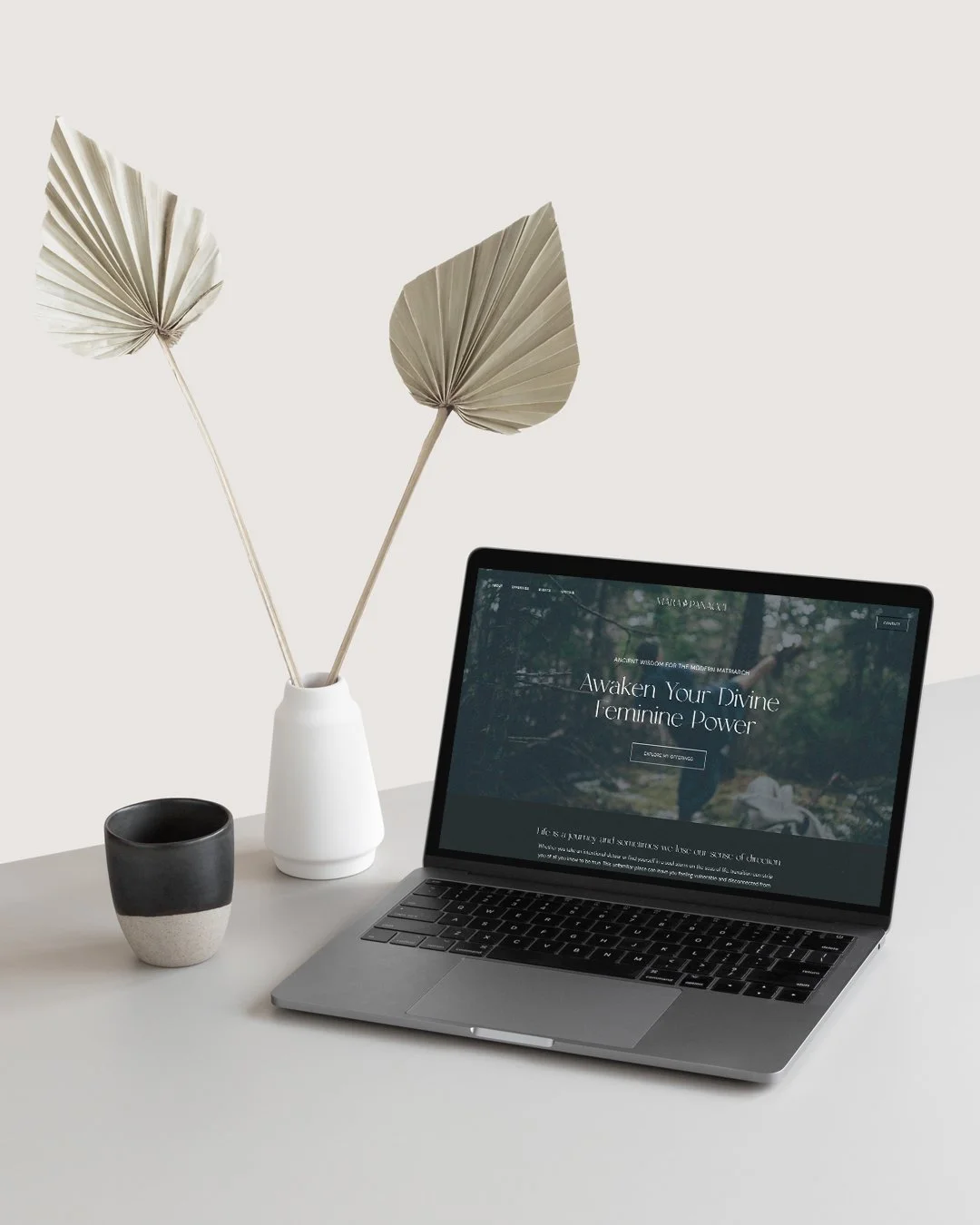 Laptop on a white desk with a website open, beside a white vase with dried palm leaves and a black and white ceramic mug.