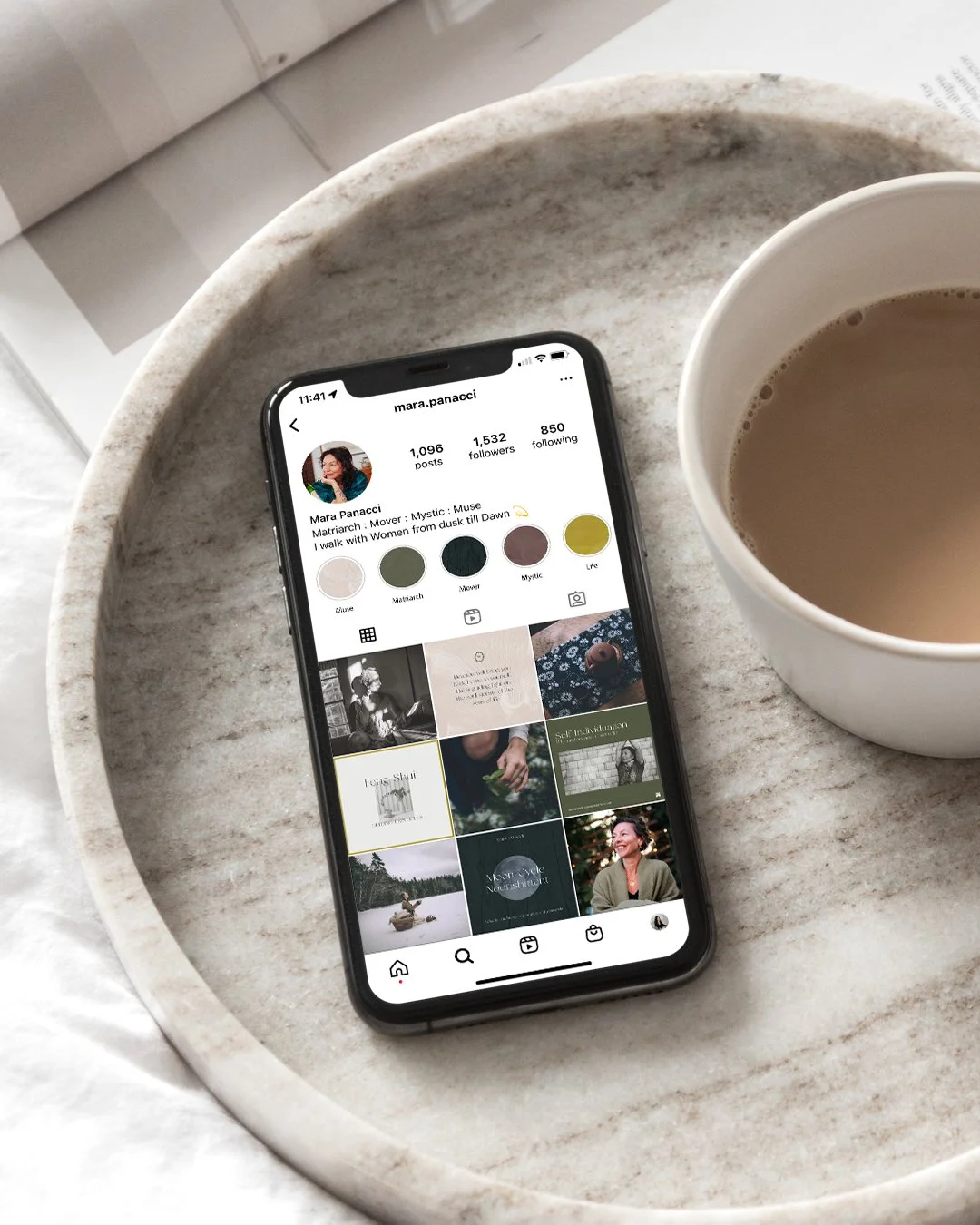 A smartphone displaying an Instagram profile page for Mara Panacci resting on a marble tray next to a cup of coffee on a gray marble surface.