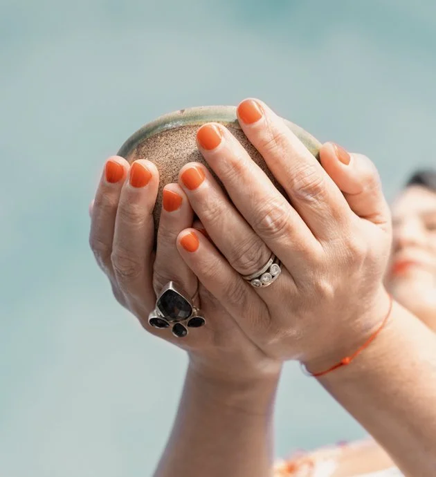 A person holding a small, round, clay object with their hands. The person has bright orange nail polish and wears multiple rings and a red bracelet.