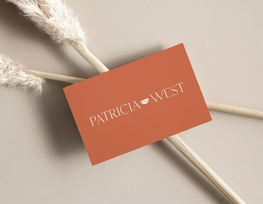 A business card with the name Patricia West on a beige background, placed on two decorative dried pampas grass stems.