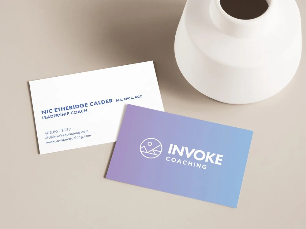 Business cards for NIC Etheridge Calder and Invoke Coaching on a beige table near a white lamp.