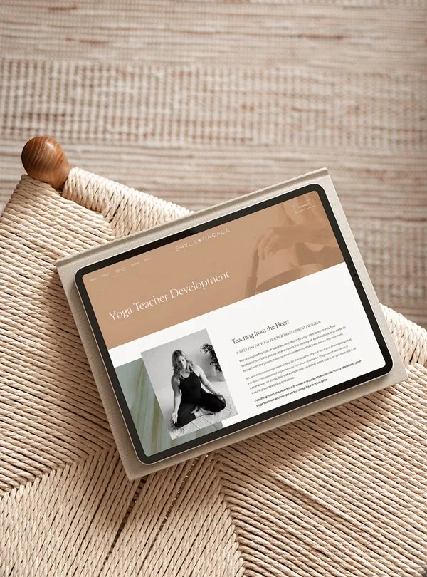 Tablet displaying a website about yoga teacher development resting on a woven chair with a wooden armrest, next to a textured wooden wall.