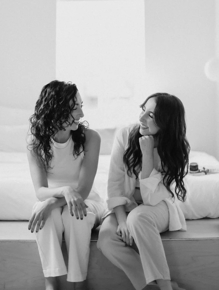 Two women sitting on a bed, smiling and looking at each other, in a cozy room.