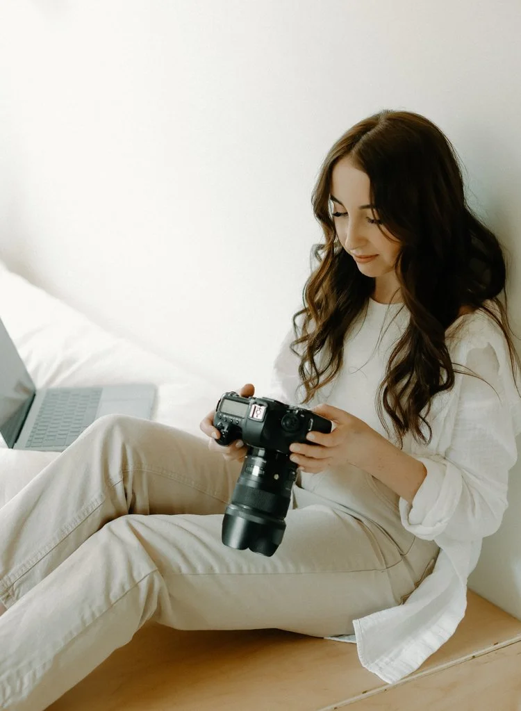 Young woman sitting on bed, holding a professional camera, smiling and looking down at it, with a laptop nearby.