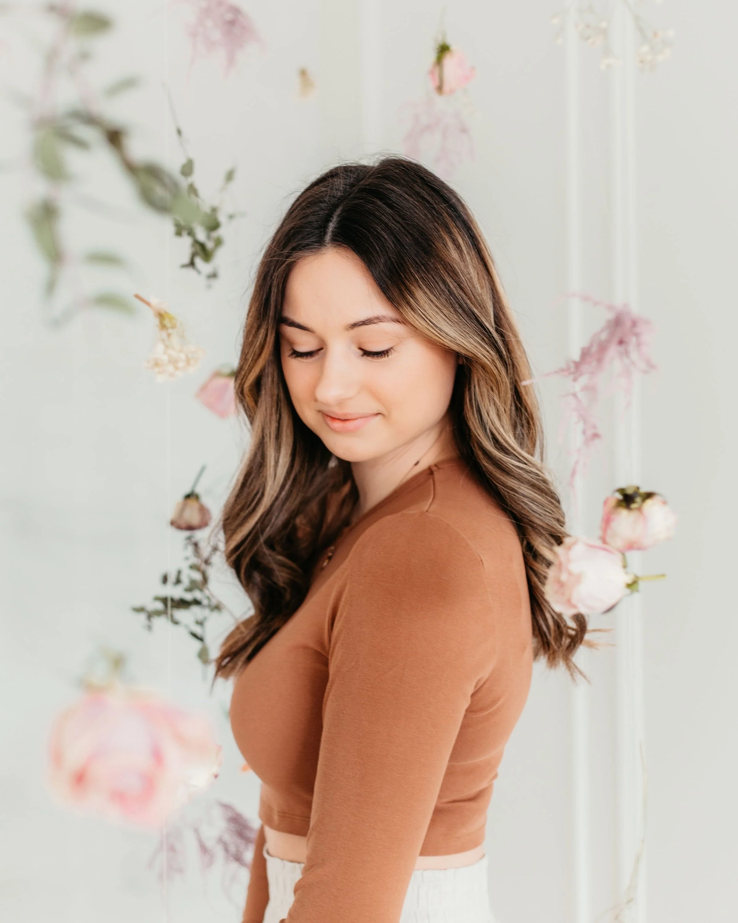 A young woman with long brown hair, wearing a tan long-sleeve top, standing with her eyes closed and a gentle smile in front of a backdrop decorated with hanging pink and white flowers.