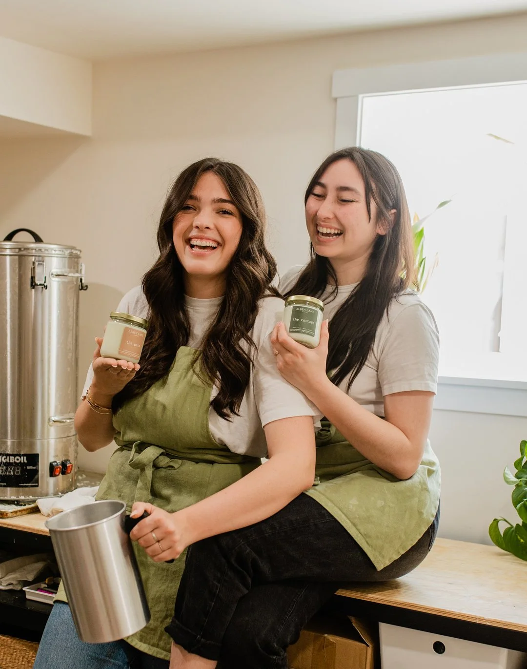 Two women in aprons sitting on a counter holding candles and laughing