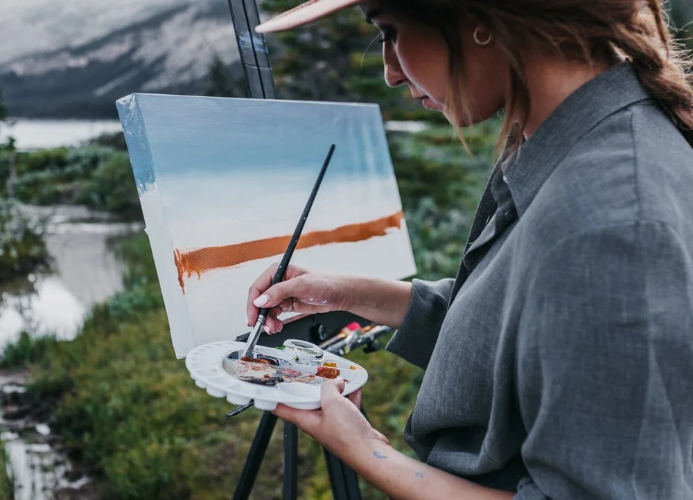 A woman painting a landscape on a canvas outdoors, with mountains and water in the background.