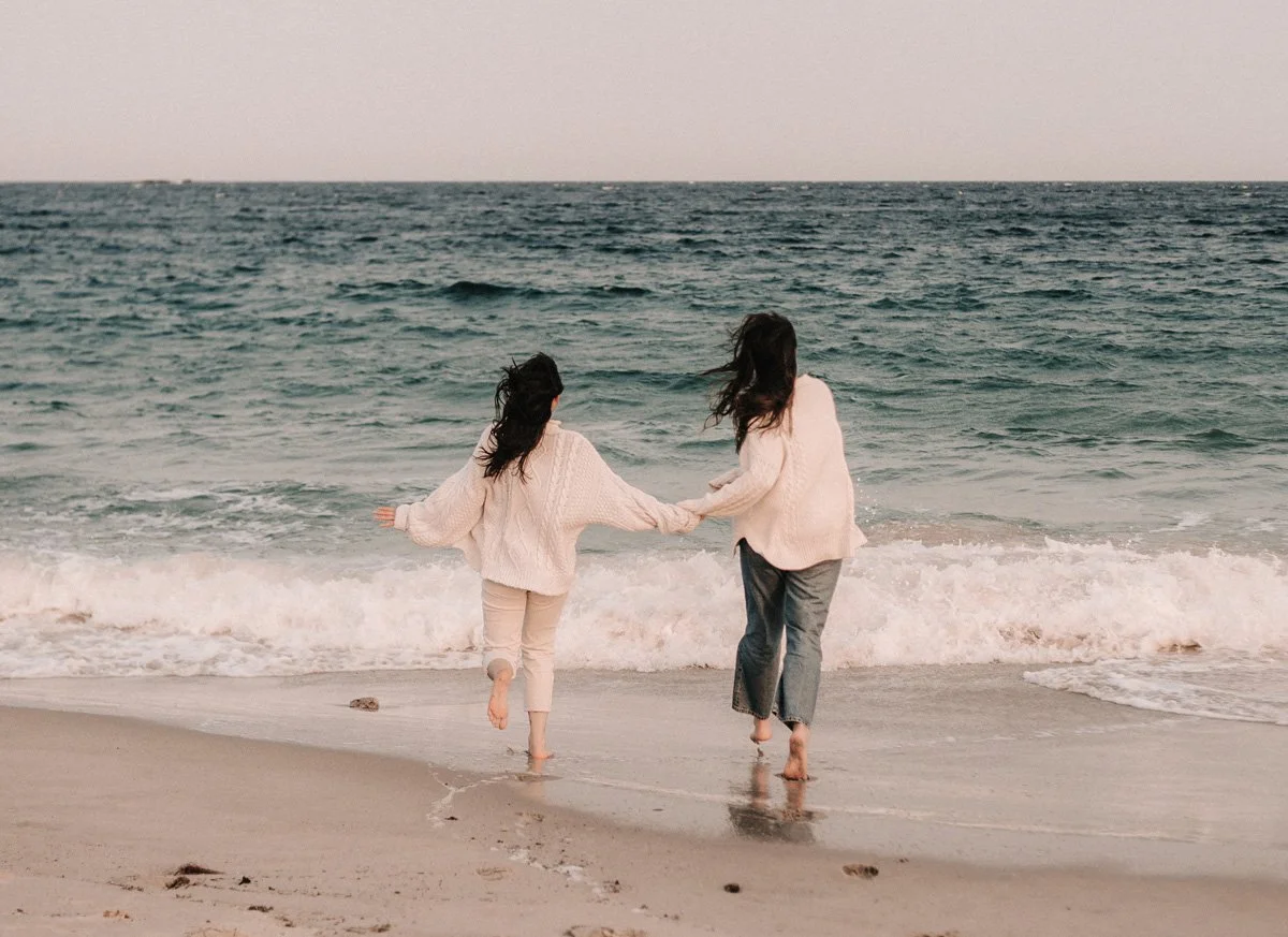 Two women walking hand-in-hand along the beach near the ocean, wearing cozy sweaters, with waves crashing on the shore.