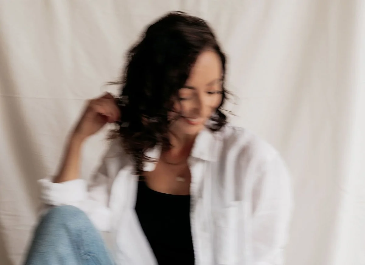 A woman with shoulder-length dark curly hair, wearing a white shirt over a black top, sitting against a neutral background.