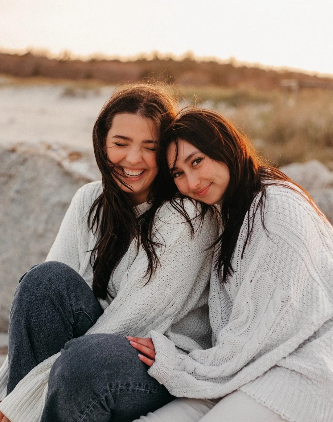 Two young women with dark hair sitting closely together outdoors at sunset, smiling and leaning their heads together, dressed in light-colored sweaters.