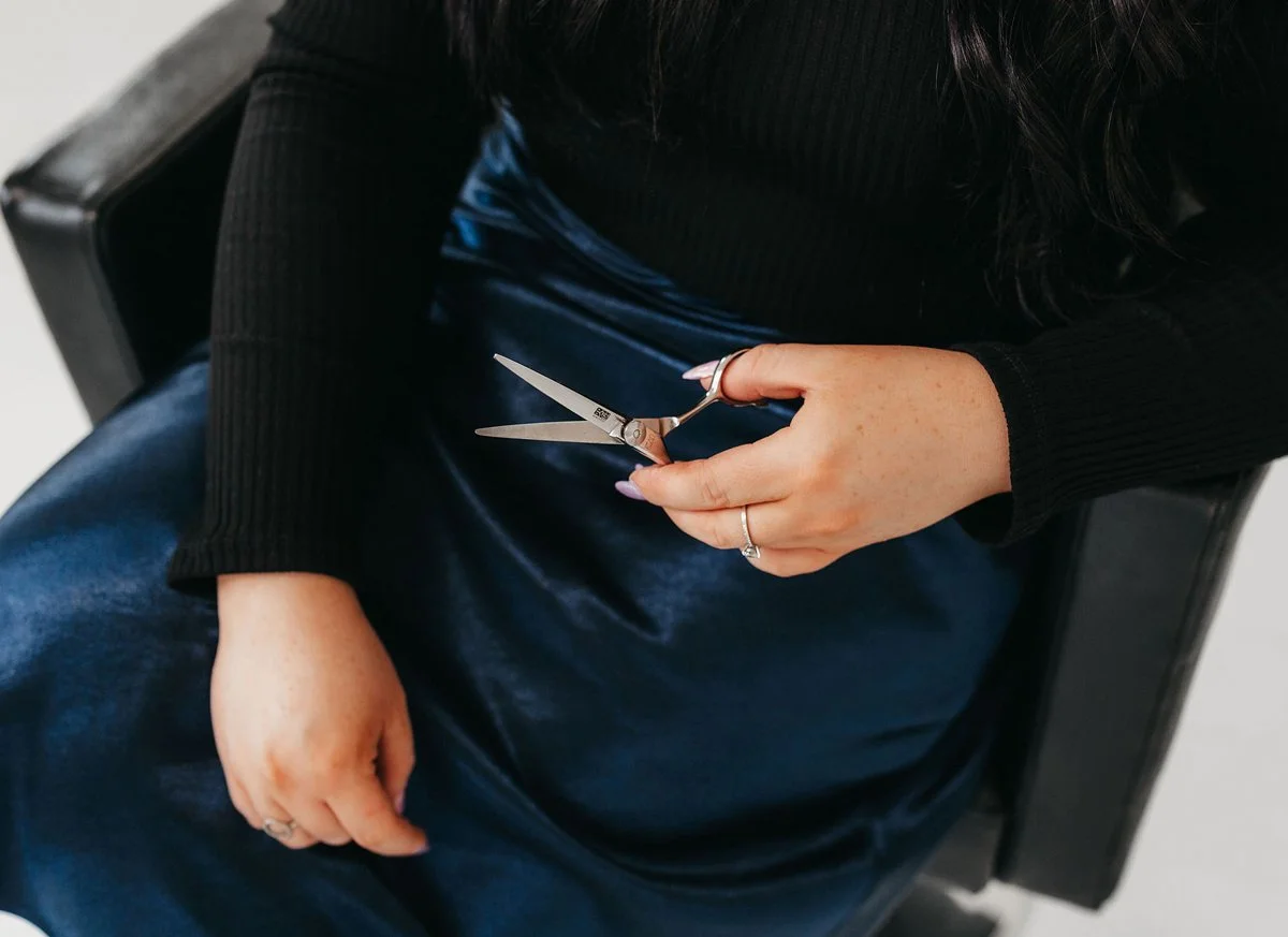 A person seated in a black armchair holding scissors in their right hand, with a manicure of polished nails and wearing rings. The person is dressed in a black long-sleeve top and a shiny blue skirt.