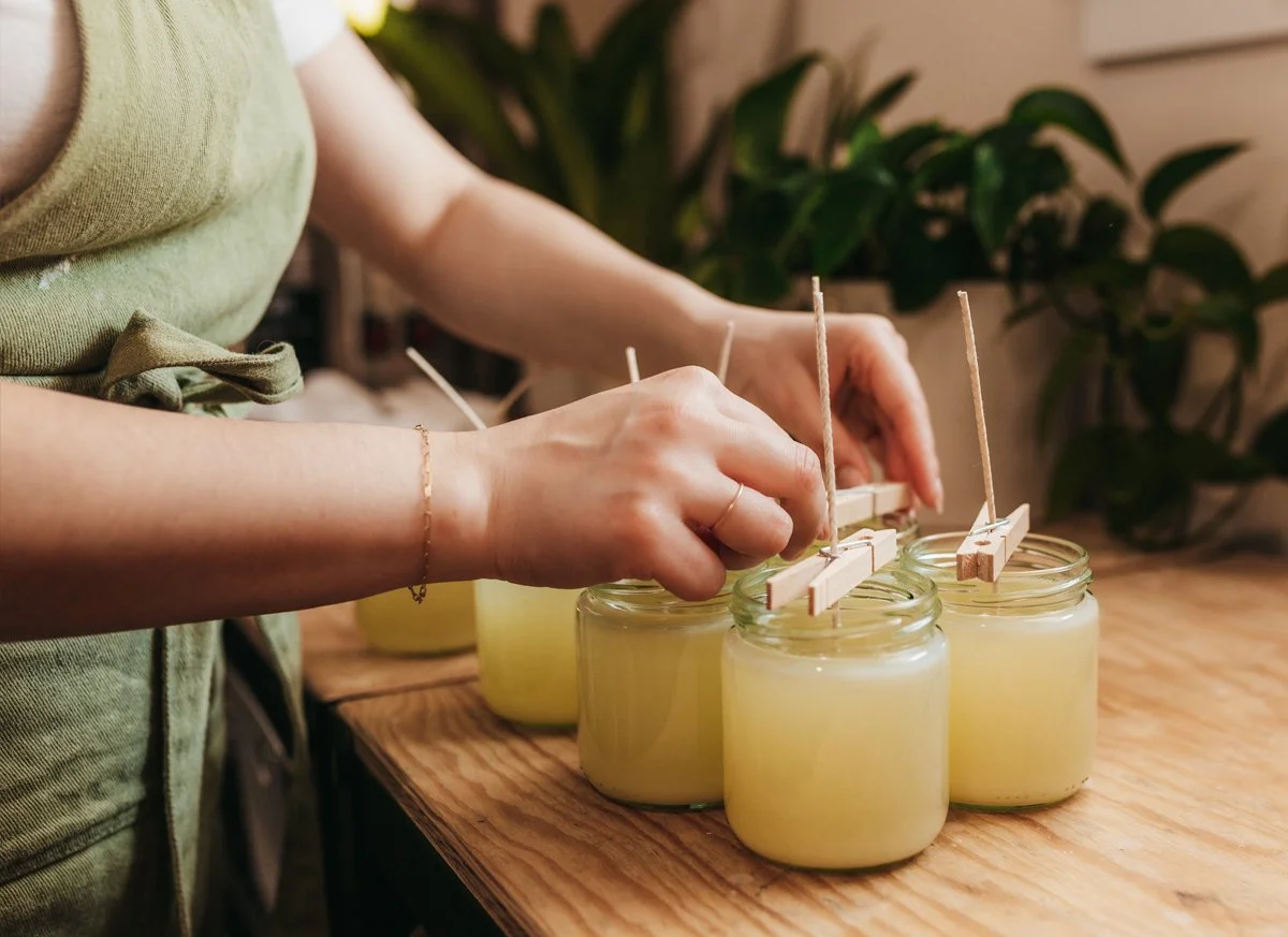 A person placing wooden wicks into jars of yellow candle wax on a wooden surface.