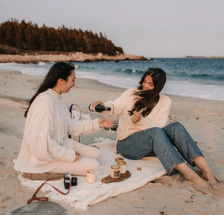 Two women having a picnic on the beach with candles and a wine bottle, one pouring wine into a glass for the other, with ocean waves and a rocky coastline in the background.