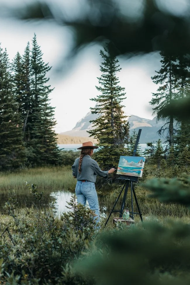 Woman painting landscape outdoors with trees, mountains, and lake in background.
