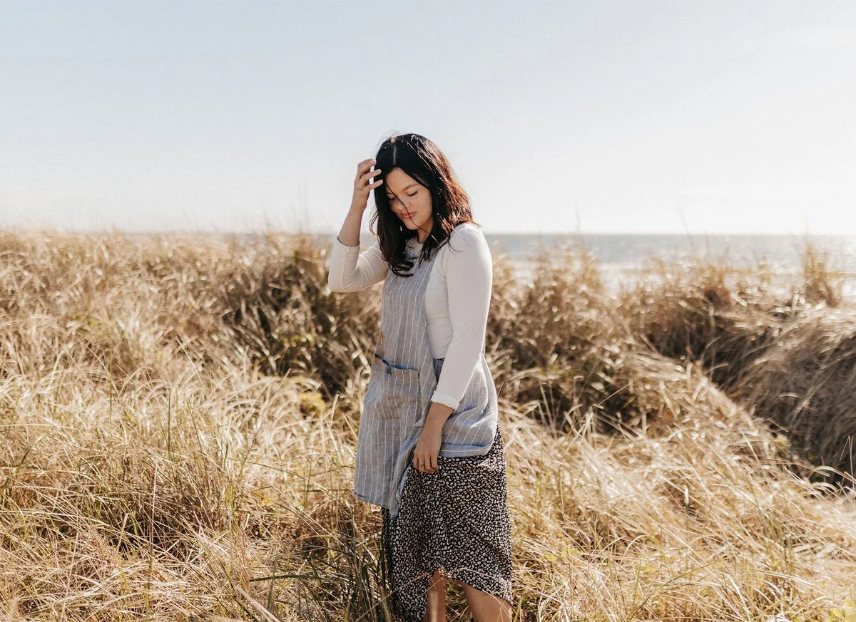 A woman standing in a field of tall, golden grass with the ocean in the background, wearing a white long-sleeve top, a gray pinafore dress, and a black and white patterned skirt.
