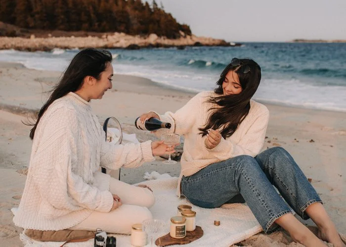 Two women sitting on a beach, smiling, as one pours a drink into the other's glass, with candles and a camera nearby.