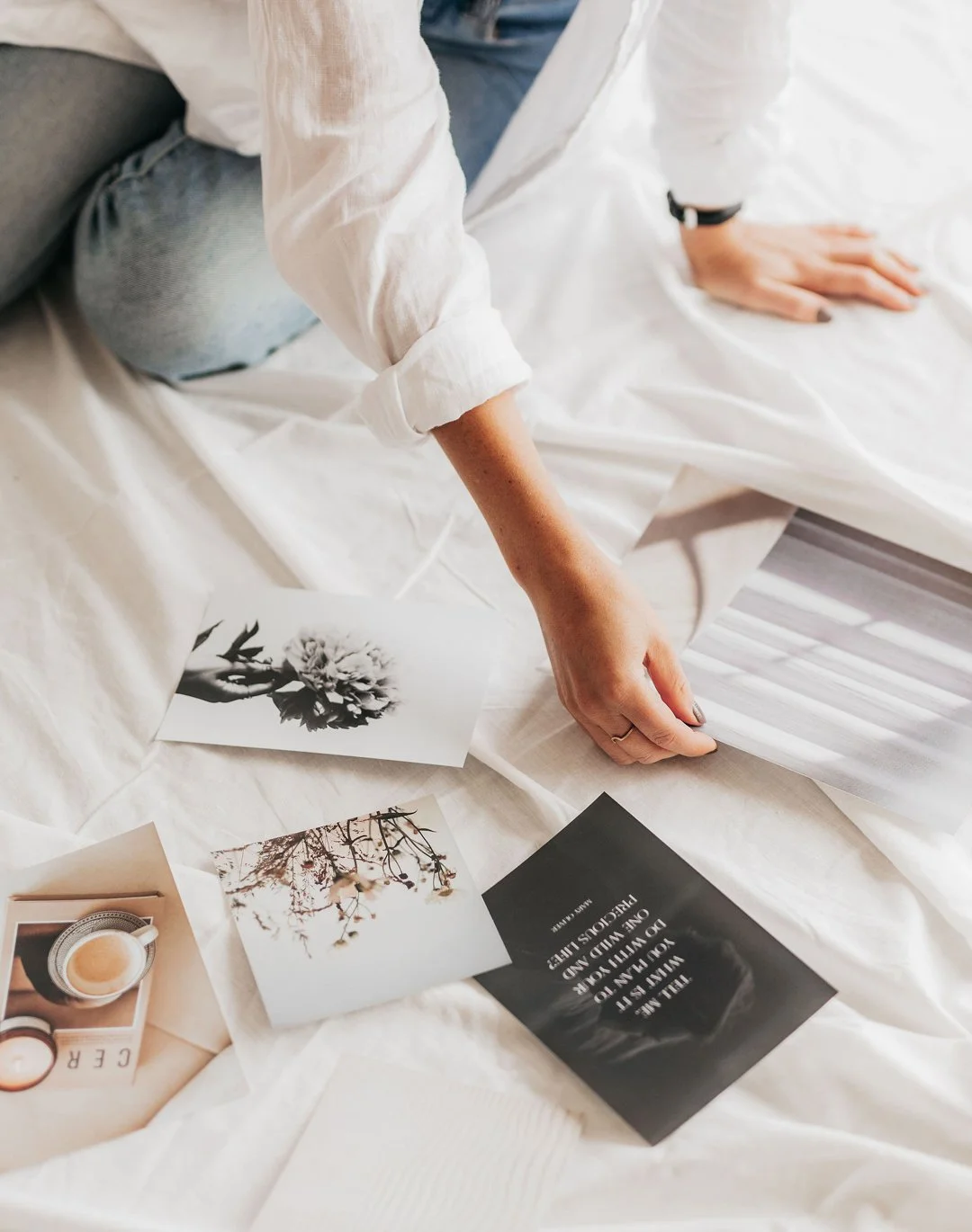Person's hand and knee on white bedspread, surrounded by black-and-white and color photographs, a card, and a cup of coffee.