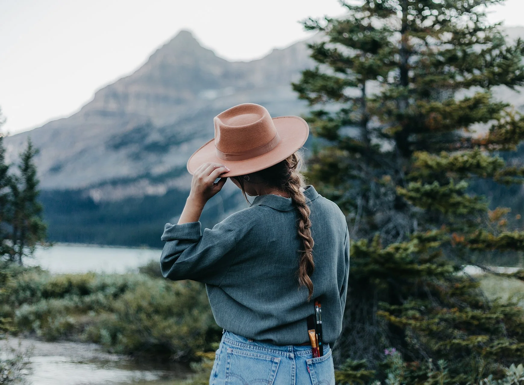 Woman with a braid in a brown hat and dark gray jacket standing in front of a mountain landscape with trees and lake.
