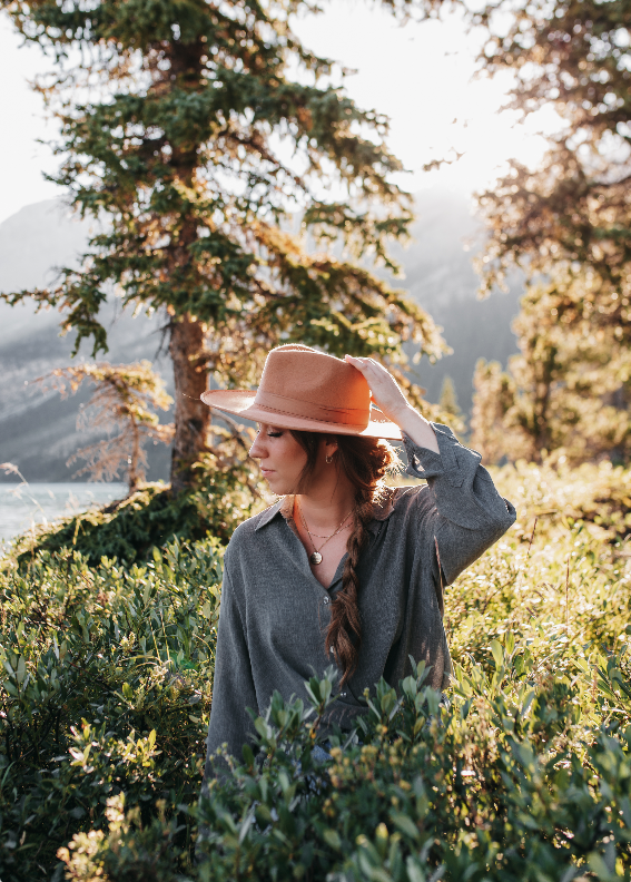 A woman wearing a large tan hat and gray shirt standing outdoors among greenery with trees and mountains in the background during sunset.