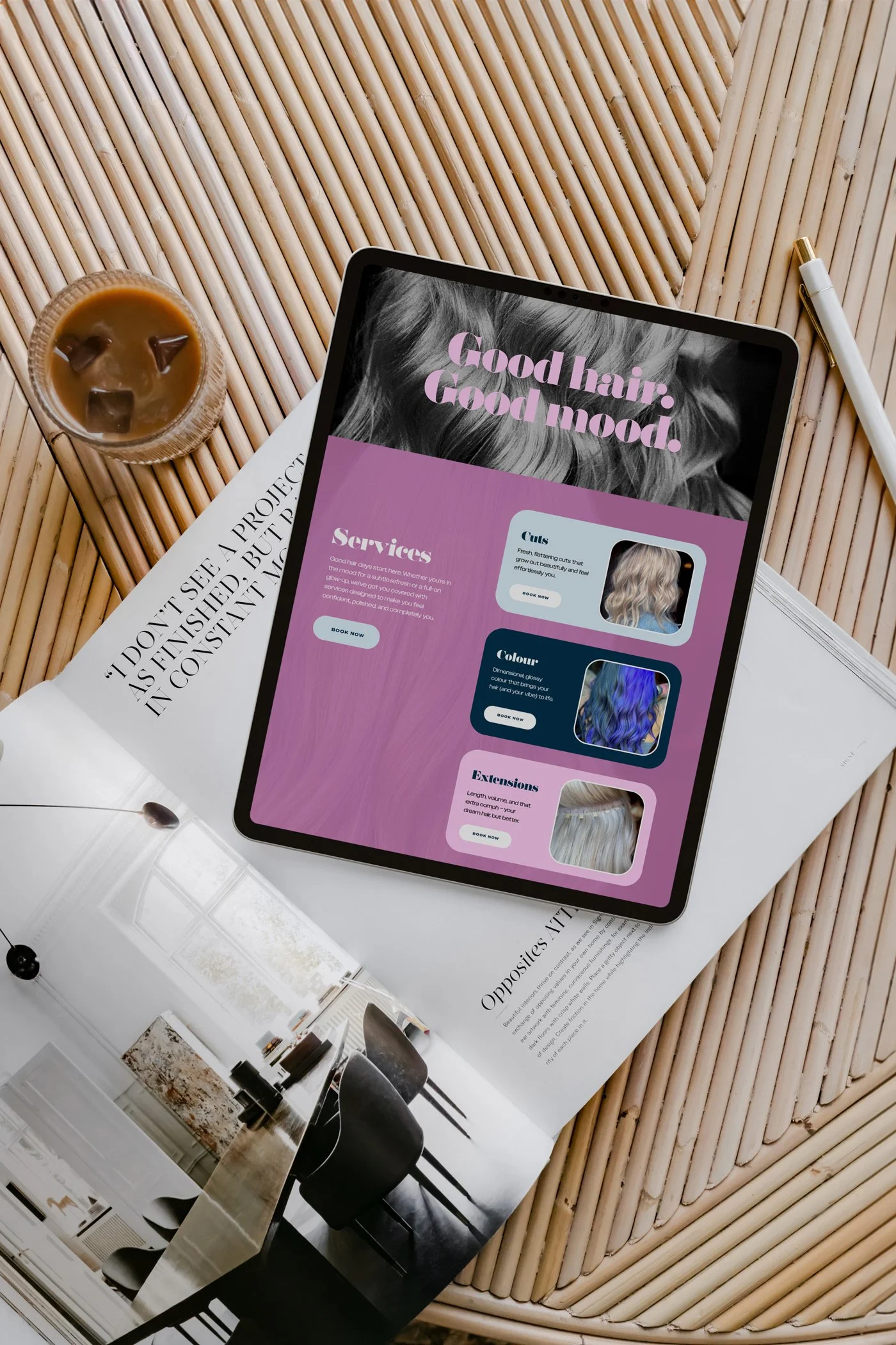 A tablet displaying a webpage about hair services, a cup of iced coffee with ice cubes, a gold and white pen, and an open magazine with interior design images are arranged on a woven rattan table.