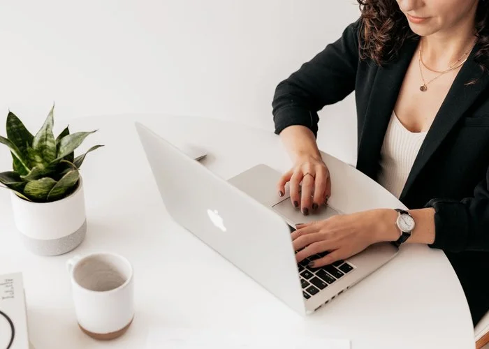 A woman working on a silver MacBook laptop at a white desk with a potted plant, a mug, and some papers.