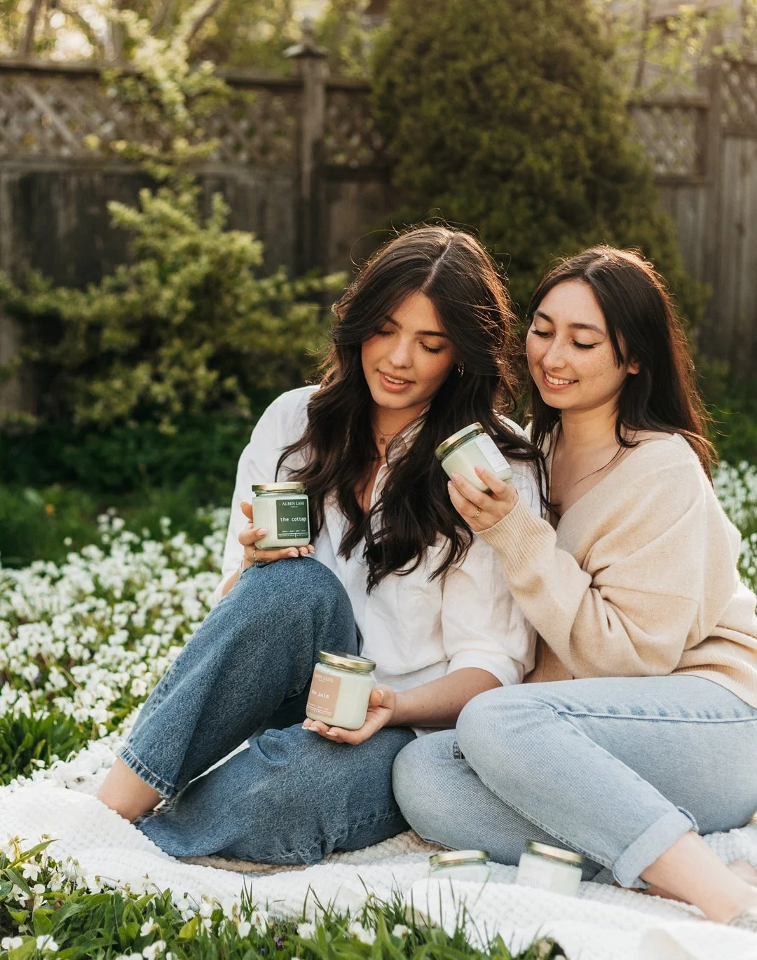 Two women sit on a blanket in a garden, holding candles or scented jars, enjoying a sunny day surrounded by greenery and flowers.
