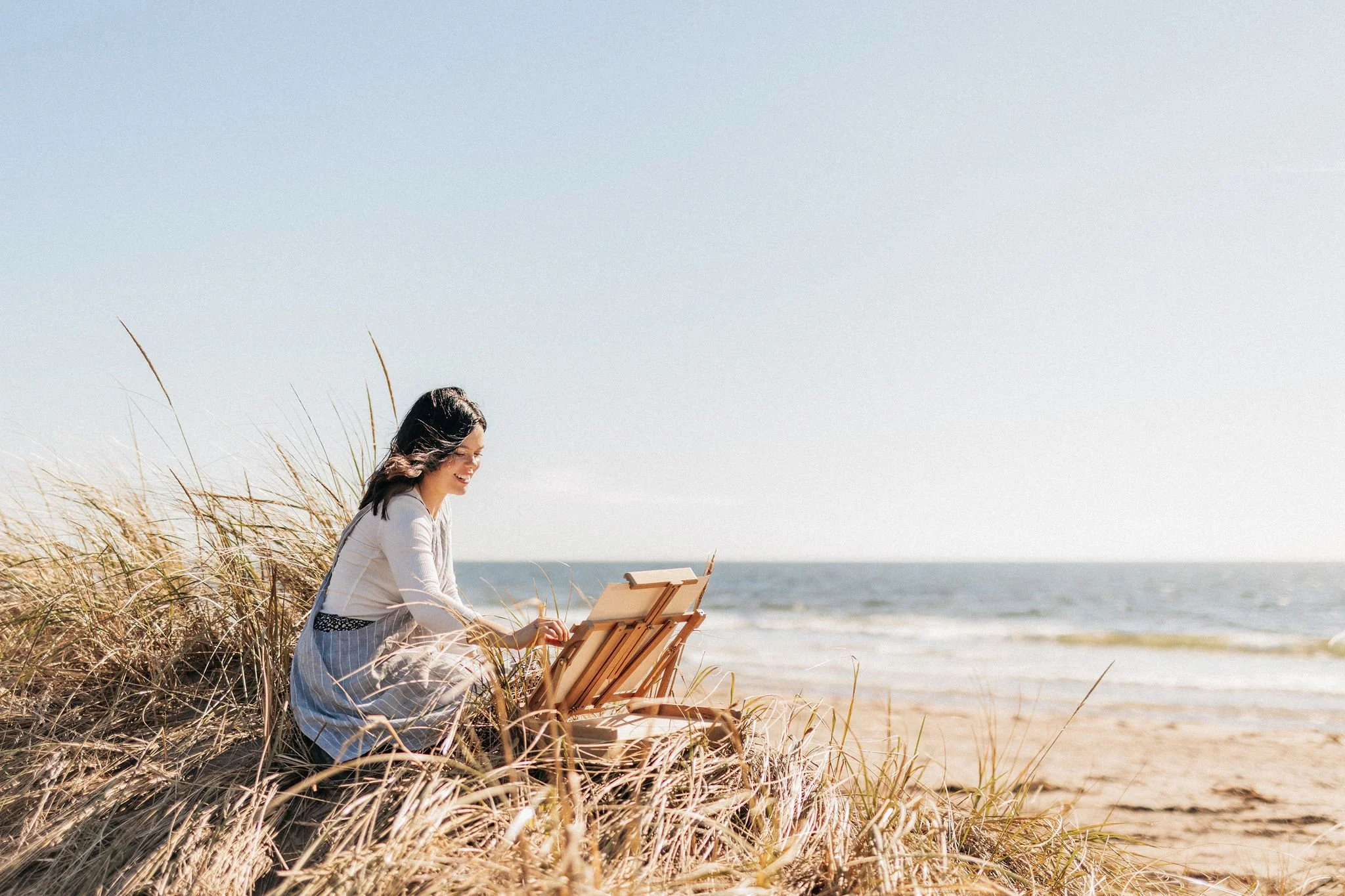 Woman sitting in sand dunes on the beach, painting on an easel with the ocean in the background.