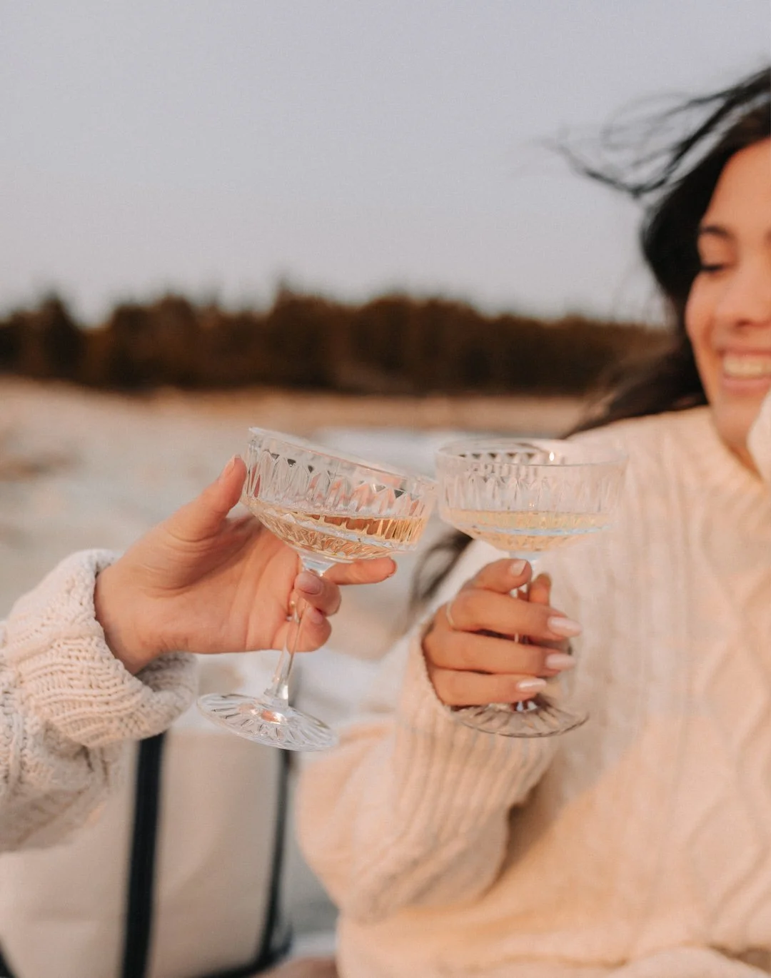 Two women clinking glasses of champagne outdoors at sunset, smiling, with a blurred background of trees and sky.