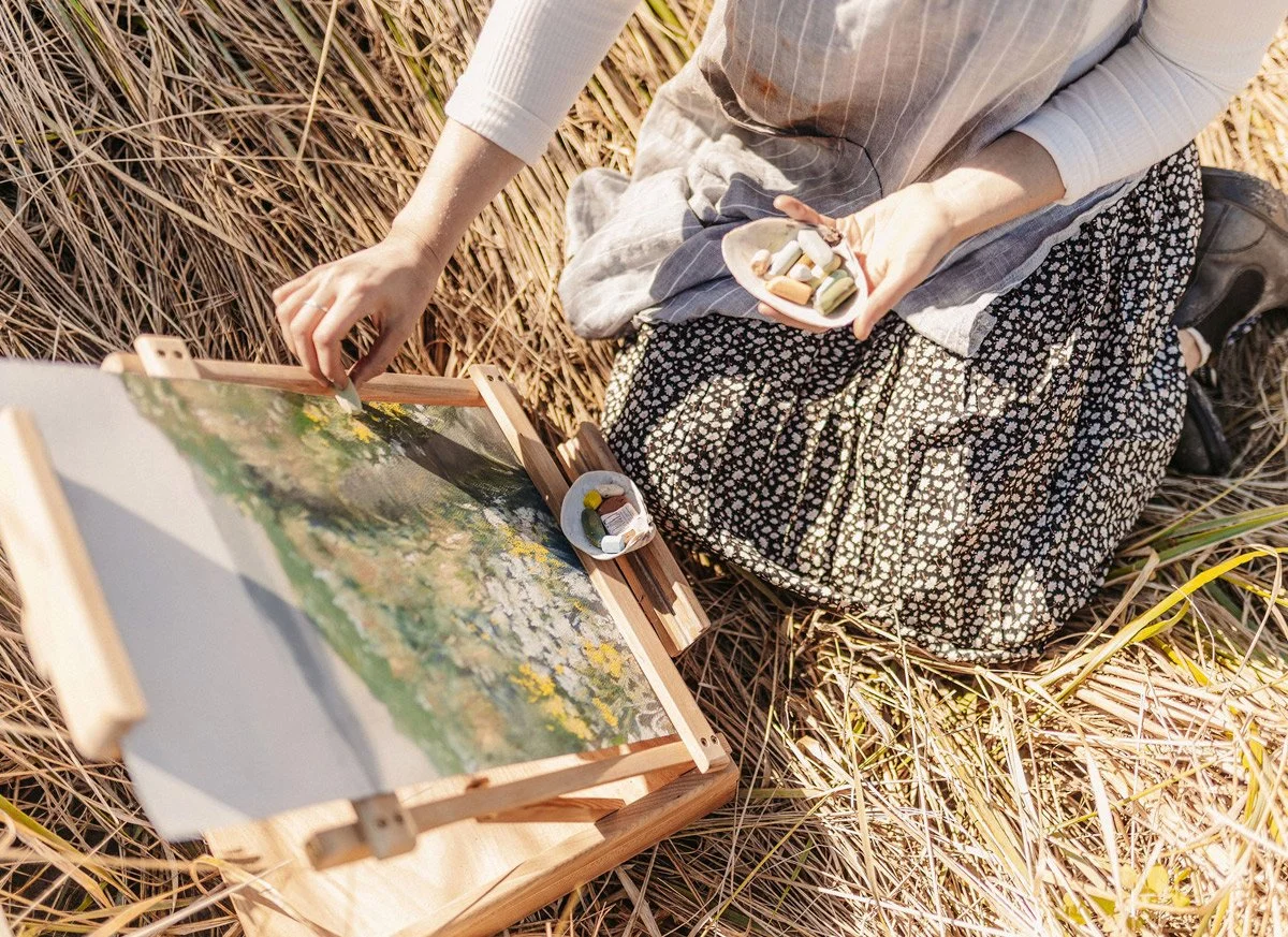 Person sitting in a field of tall grass, painting on an easel with a palette of paints, with small bowls of paint nearby.