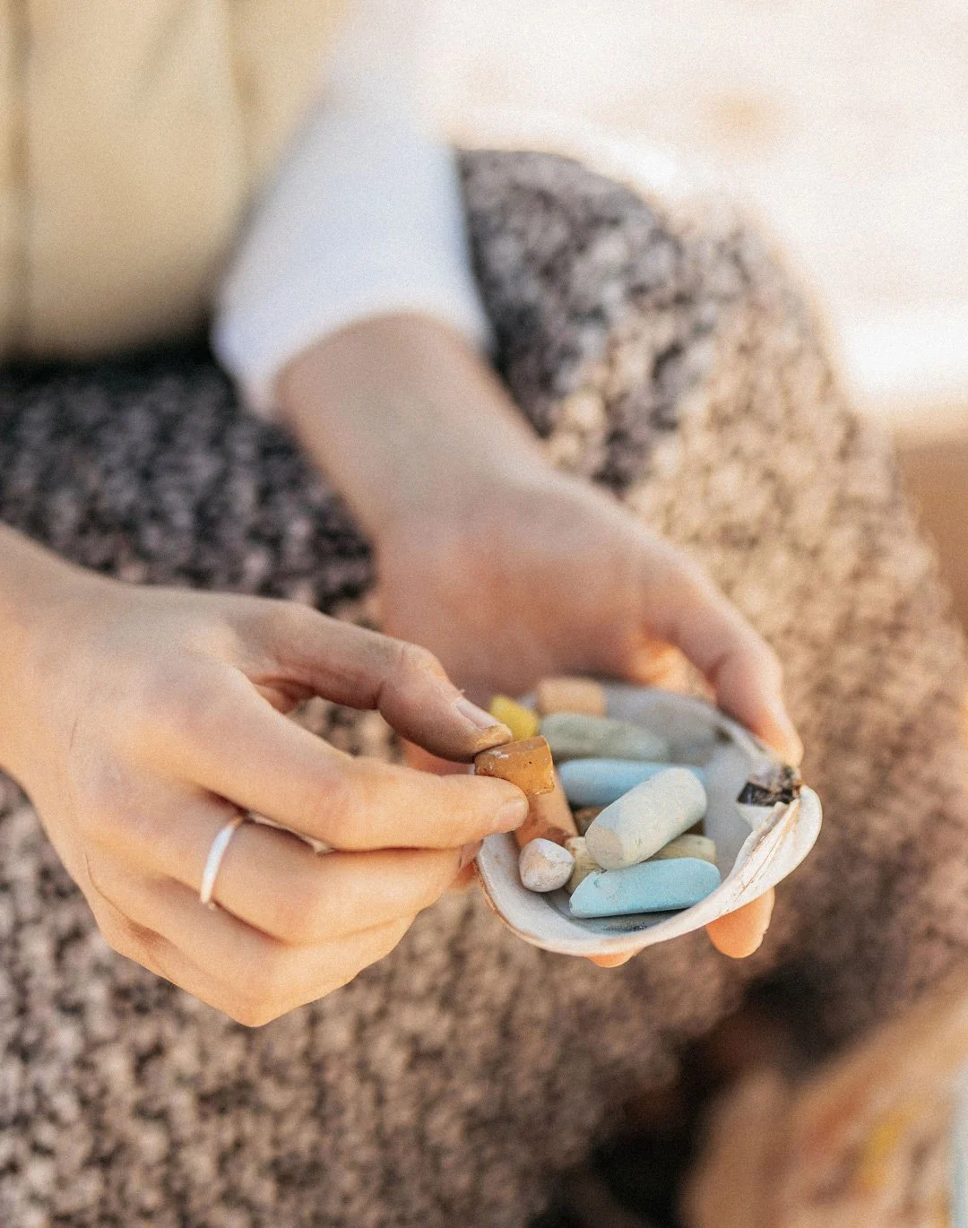 Person holding a small dish filled with pastels, wearing a light-colored top and sitting on a carpeted floor.
