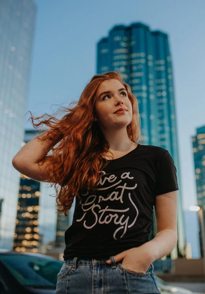 A young woman with long red hair, wearing a black T-shirt with the text 'Have a Great Story' and jeans, stands outdoors in an urban setting with tall skyscrapers in the background, during dusk.