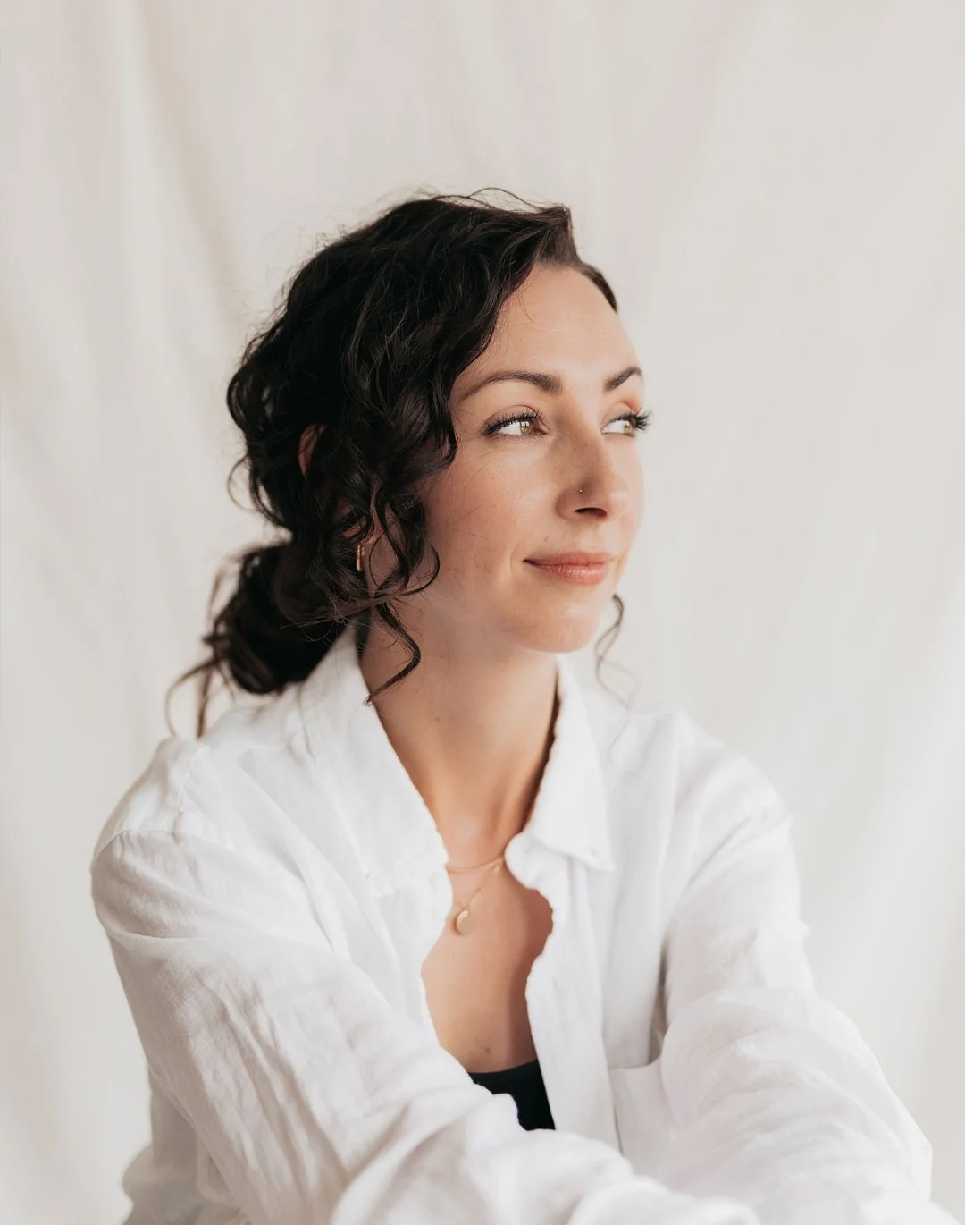 A woman with dark curly hair and light skin, wearing a white shirt and a moon-shaped necklace, looking sideways with a slight smile.