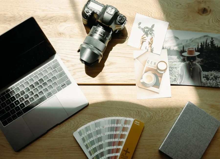 A wooden desk with a laptop, a professional camera with a lens, printed photographs, a color swatch fan deck, and a gray notebook.