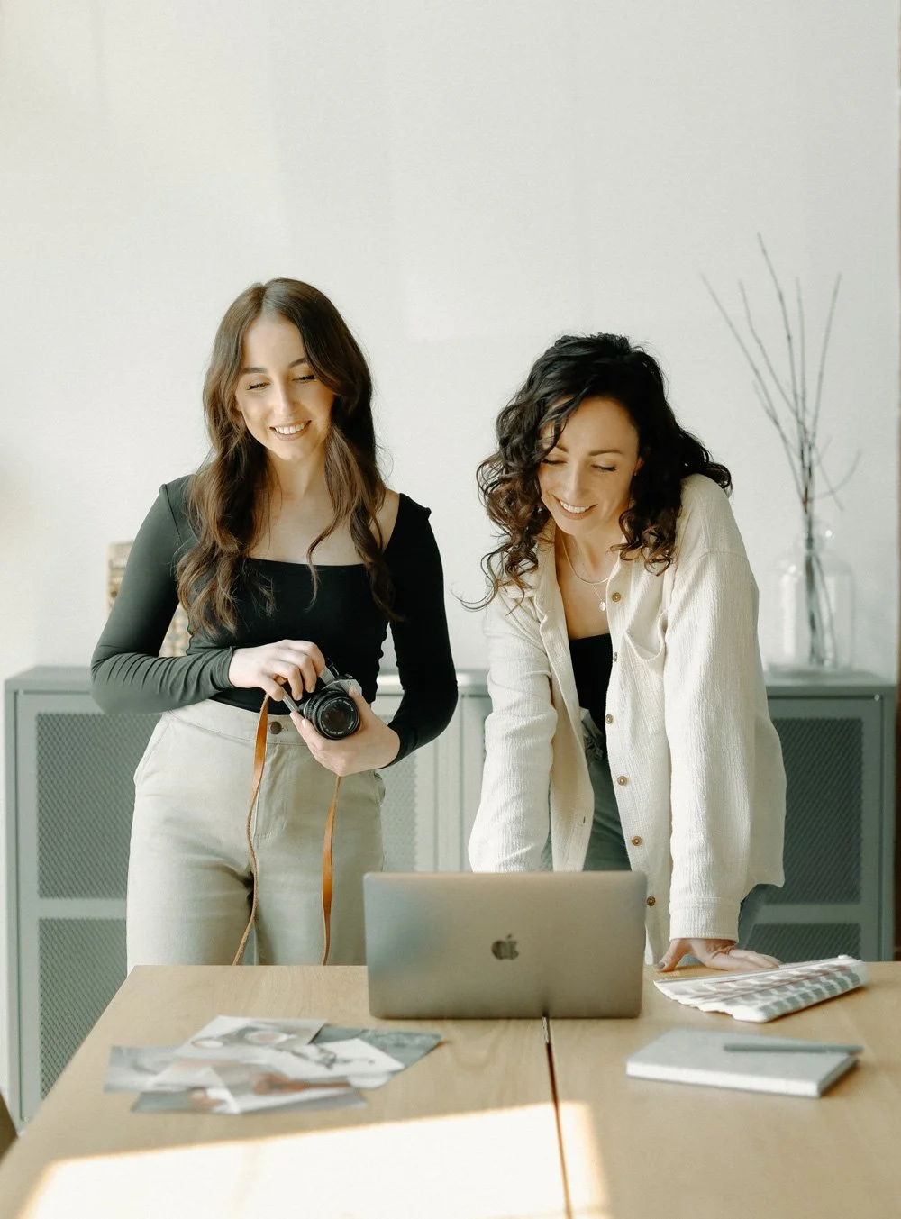 Two women collaborating on a project in an office, one holding a camera and the other looking at a laptop.