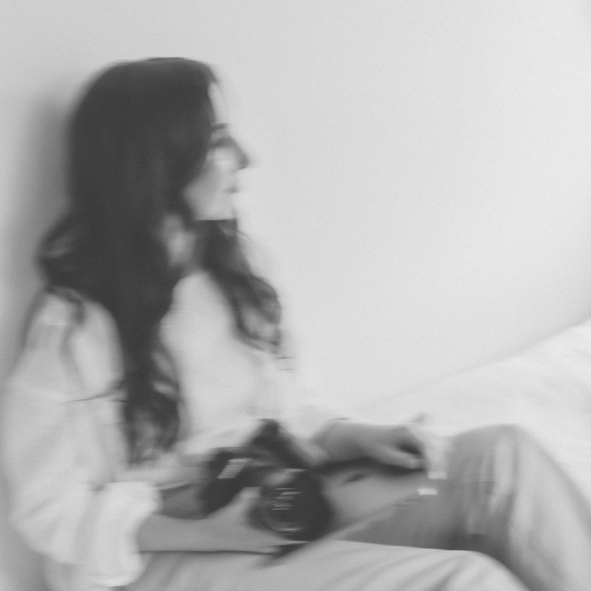 A woman with long dark hair sitting on a bed, holding a camera in her hands, looking to the side, in a monochrome photo.
