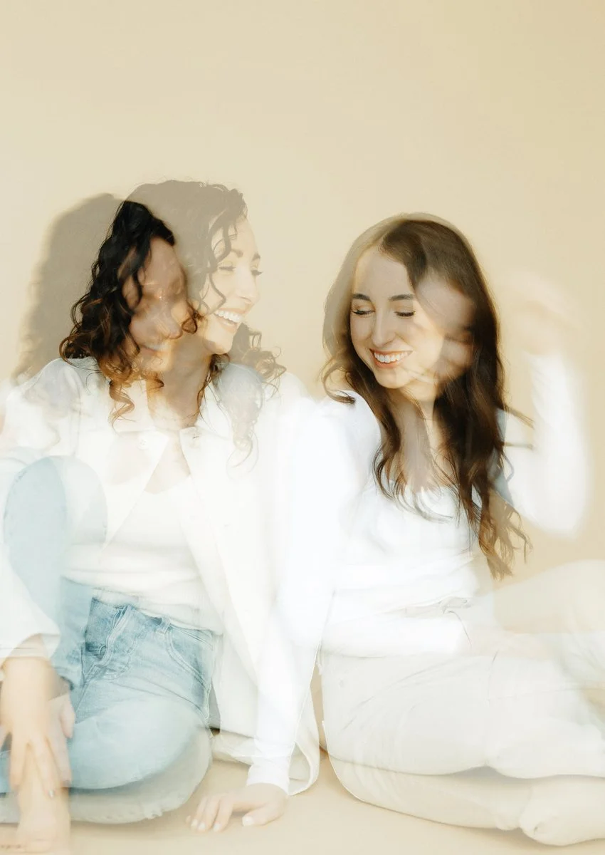 Two women sitting on the floor, smiling and appearing to be in motion, with a double exposure effect creating overlapping images of them.