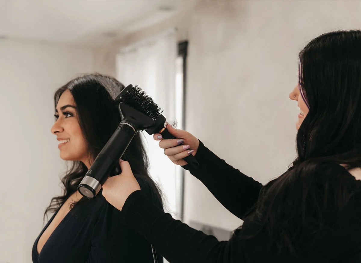 A woman with long dark hair is getting her hair dried with a handheld blow dryer by a stylist in a room with natural light.