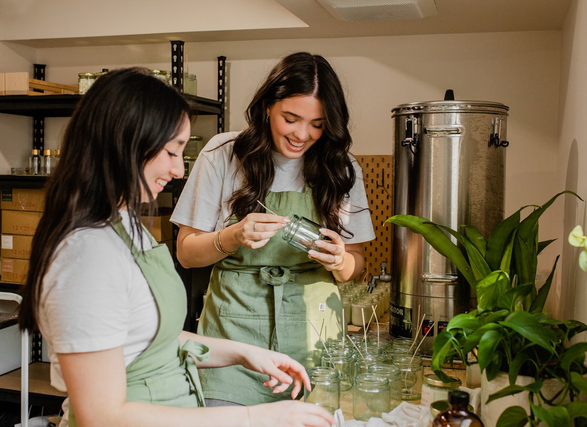 Two women in aprons preparing jars in a kitchen with shelves, plants, and a large stainless steel container.