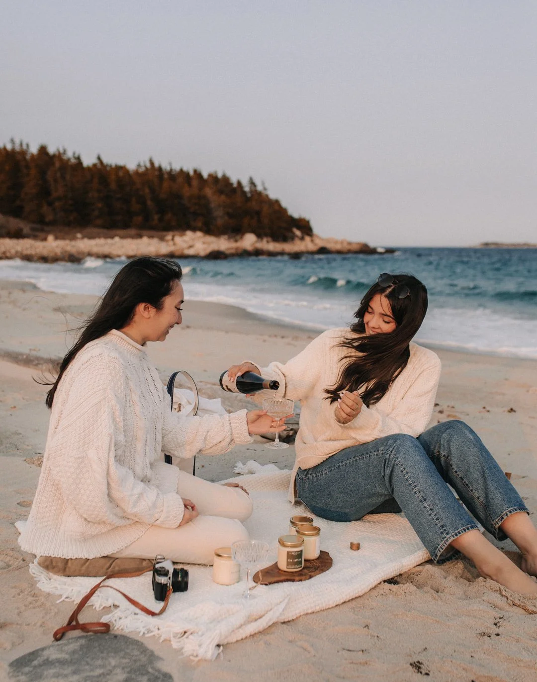 Two women having a picnic on the beach, one is pouring drinks into a glass for the other, surrounded by candles and a camera, with an ocean, rocks, and trees in the background.