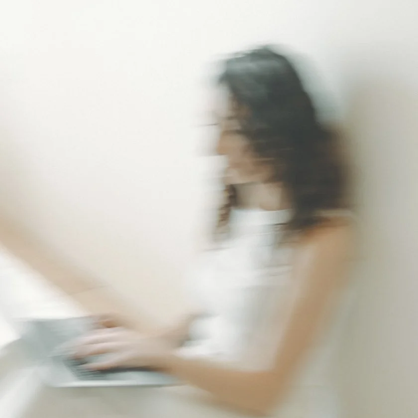 Blurry image of a young woman with dark hair working on a laptop in a minimalistic room with a white wall.