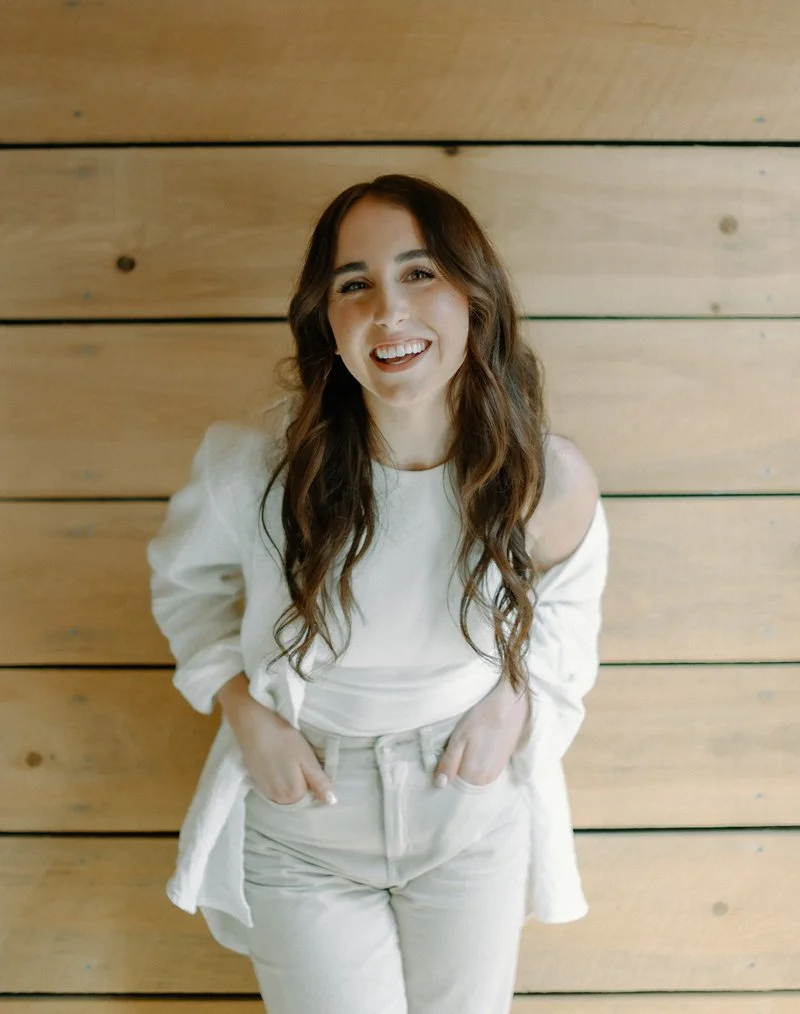 A young woman with long wavy brown hair smiling with her hands in her white pants pockets, wearing a white off-shoulder top, standing in front of a wooden wall.