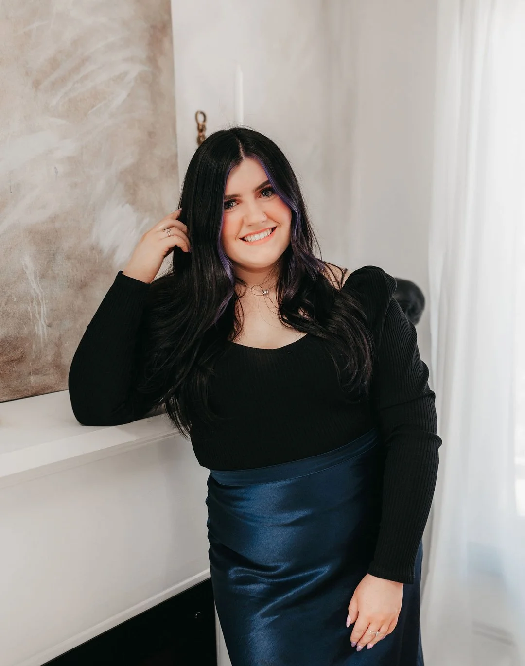 A young woman with long black hair and purple highlights, smiling, dressed in a black top and a dark blue satin skirt, standing indoors against a wall with abstract art.