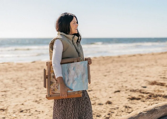 Woman standing on the beach holding an easel and canvas, looking towards the ocean on a sunny day.