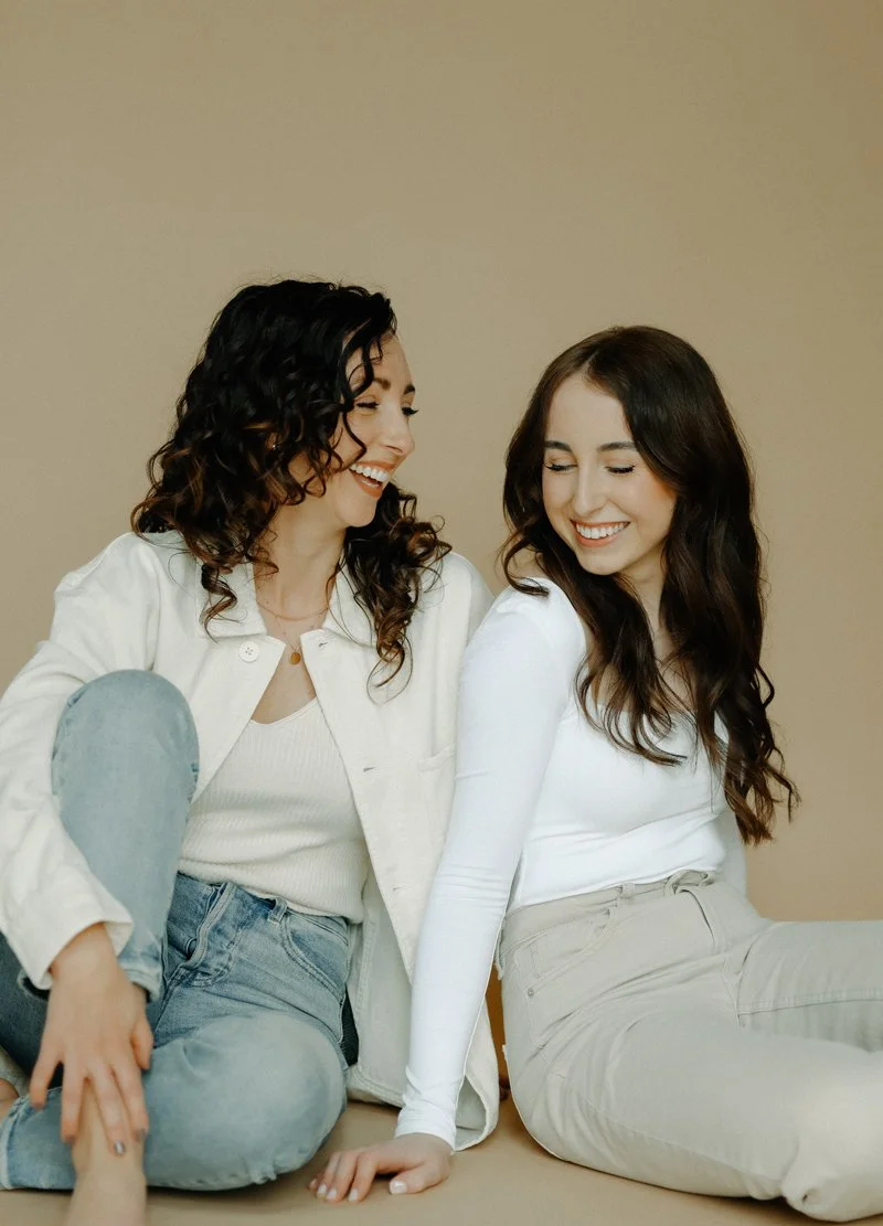 Two women sitting on the floor, smiling and engaging with each other, against a plain beige background.