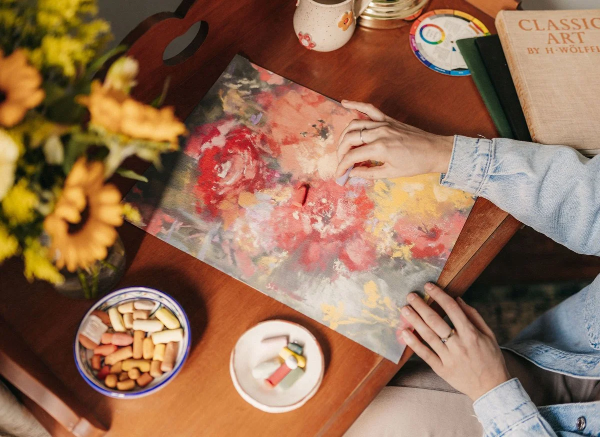 Person creating an abstract floral painting on paper, surrounded by books, a vase with flowers, and art supplies on a wooden table.