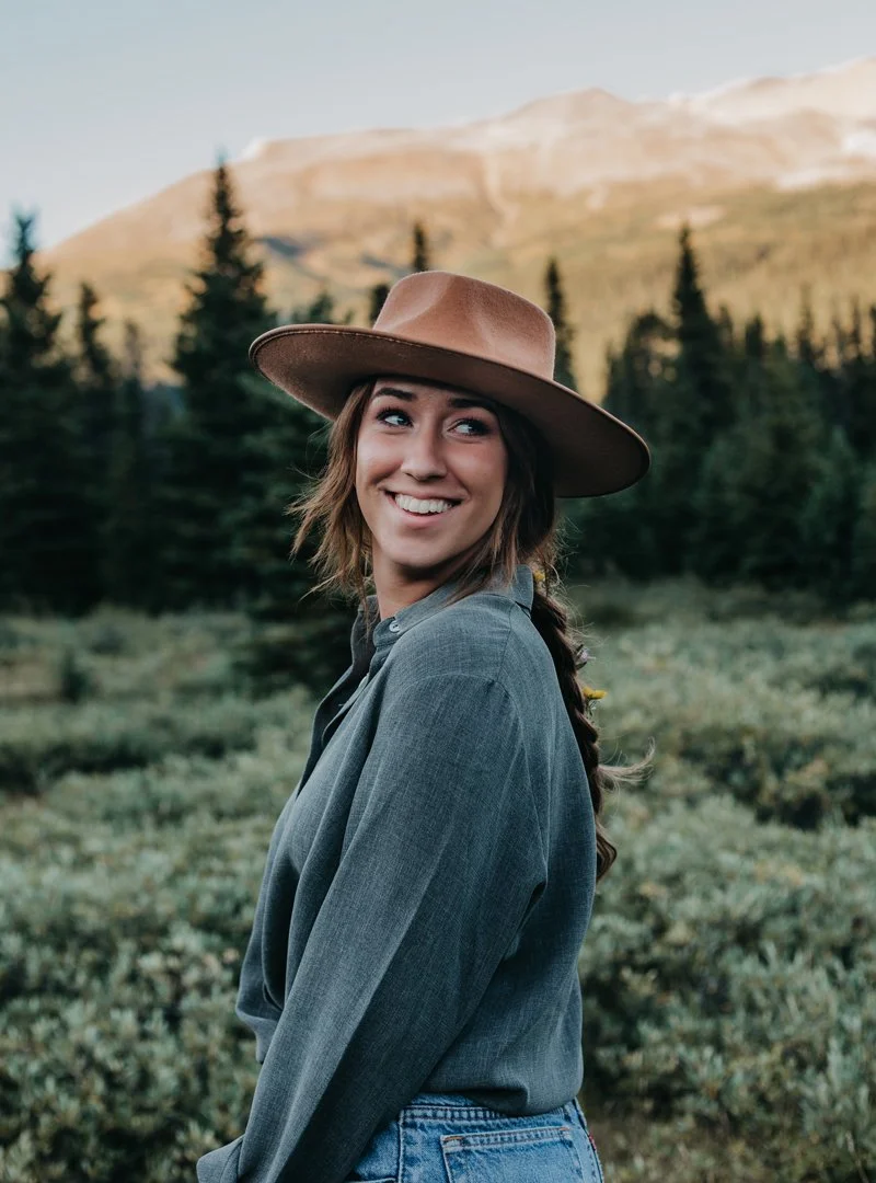 Young woman smiling outdoors in a mountain and forest setting, wearing a wide-brimmed hat and denim shirt.