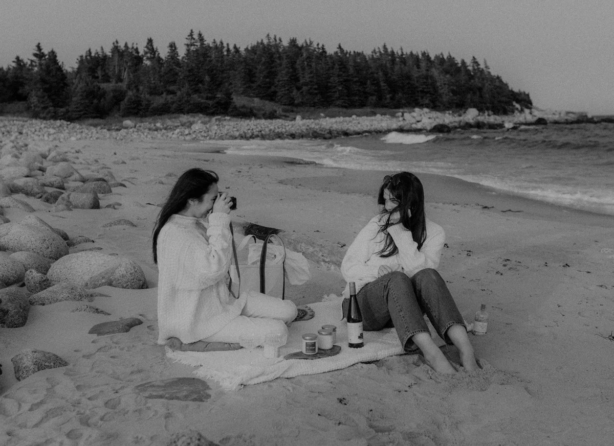 Two women sitting on a blanket on a beach, with one taking a photo of the other. There are bottles and jars on the blanket, with a scenic background of trees and rocks by the shoreline.