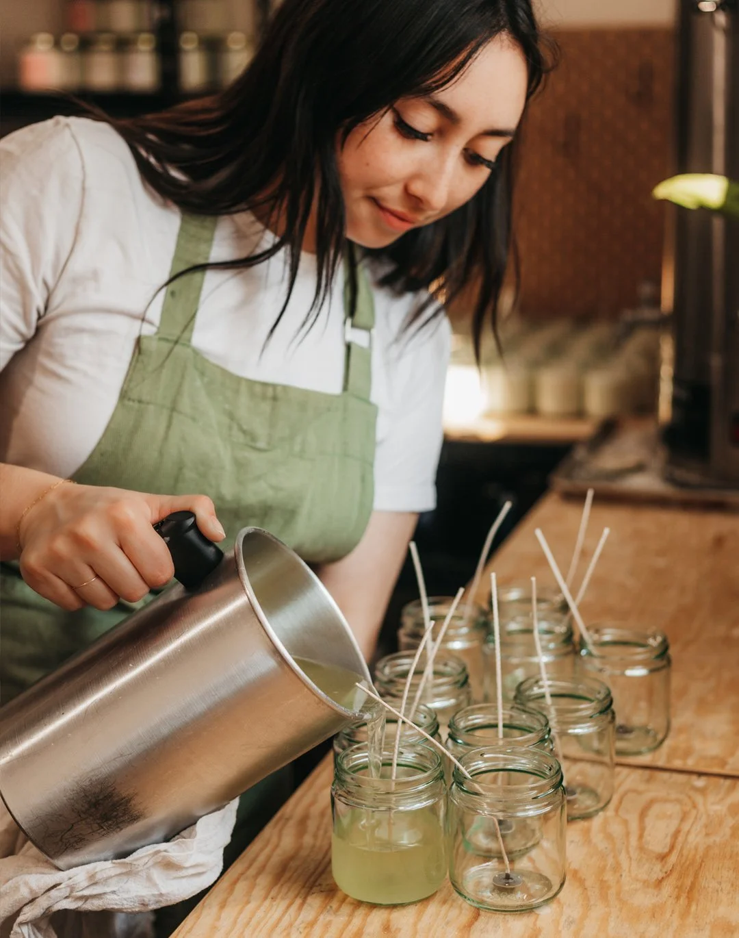 A woman wearing a green apron pours liquid wax into glass jars on a wooden surface.